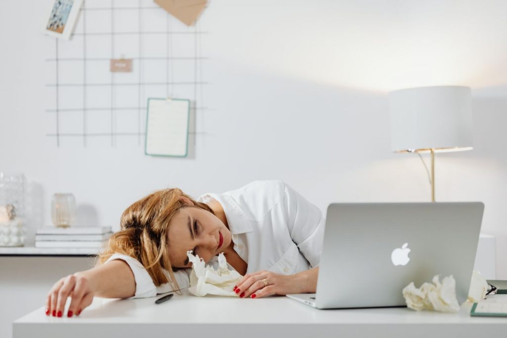 Home Exhausted woman in white attire rests head on desk beside laptop and crumpled papers.